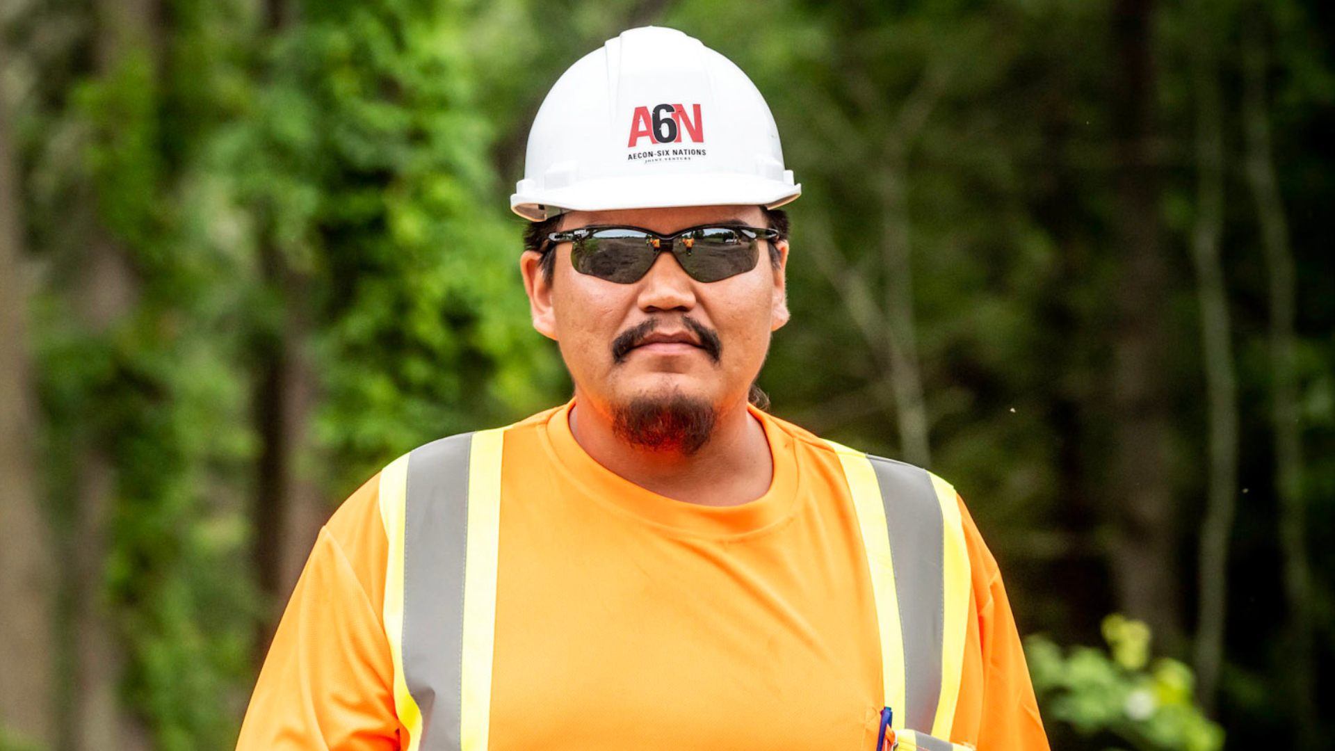 Man wearing a white hardhat and sunglasses in front of trees